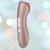 A beige, handheld Satisfyer Pro 2+ Air Pulse Stimulator + Vibration by Satisfyer, featuring a white applicator tip, is displayed against a blurred, pastel-colored background with circular light bokeh effects. The device boasts an ergonomic design with visible buttons for operation.